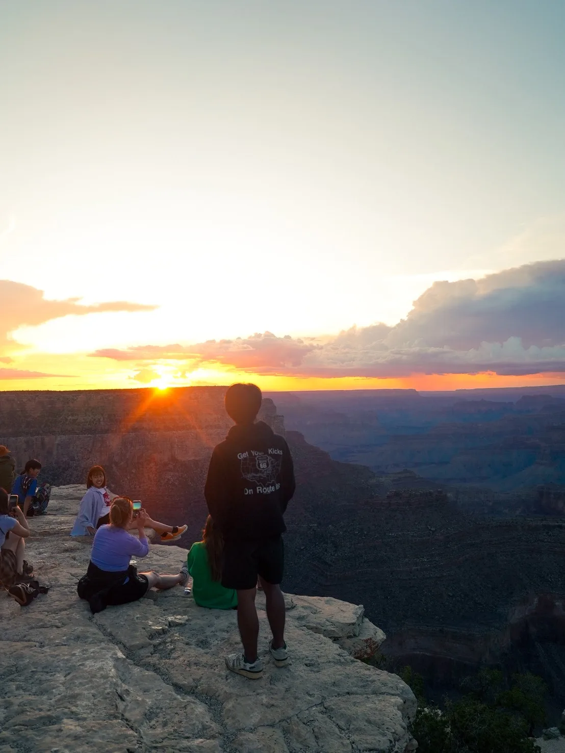 「Get Your Kicks On Route 66」のパーカーを着て、グランドキャニオンの夕日を眺める様子
