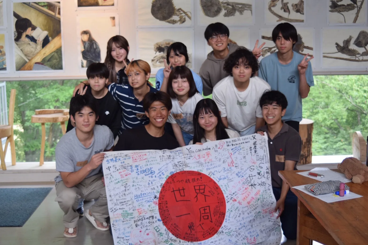 「世界一周」の​寄せ​書き旗を​囲んで​記念撮影する​参加者たち