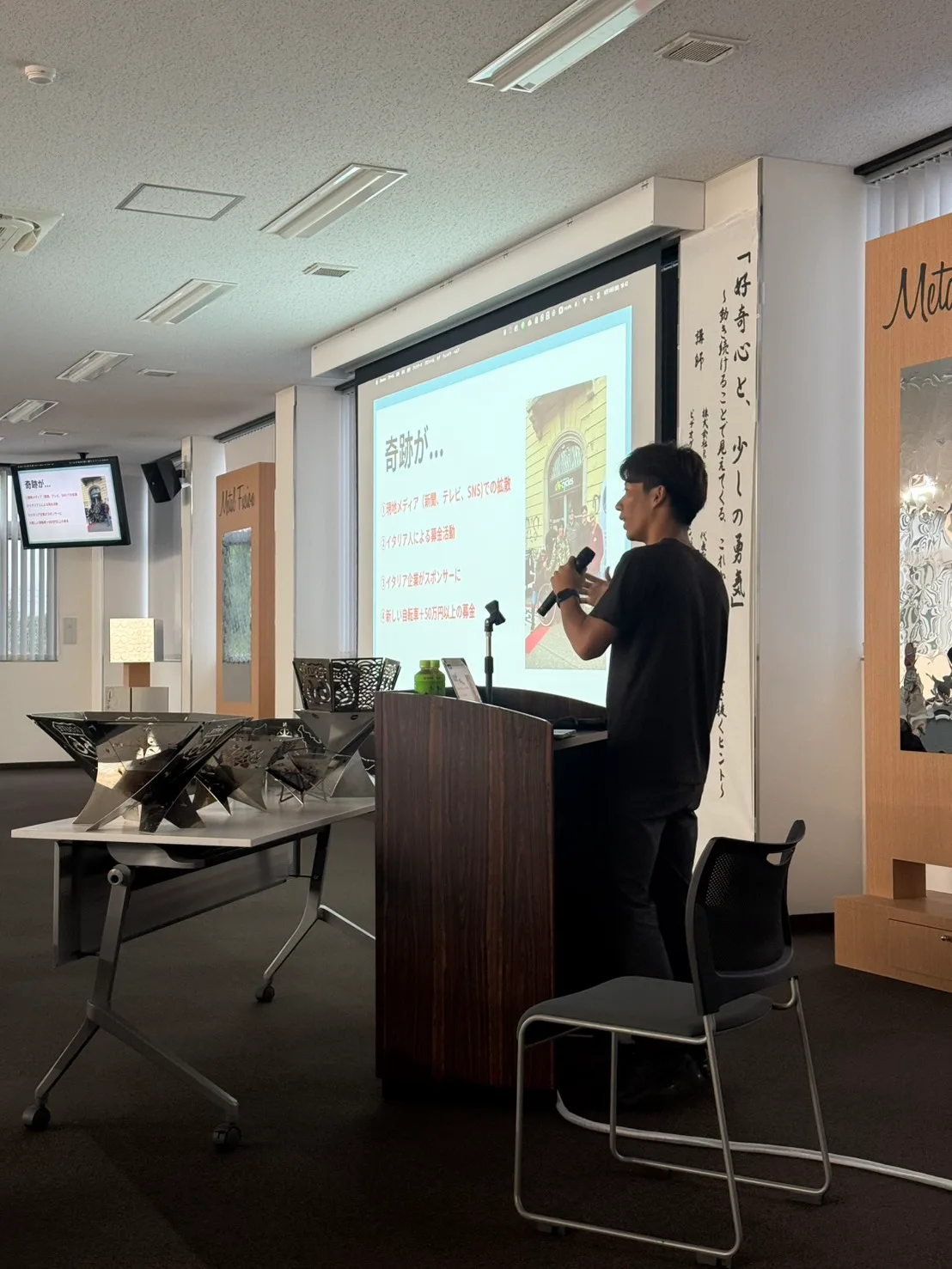 スクリーンの​前で​スライドを​使って​話す登壇者