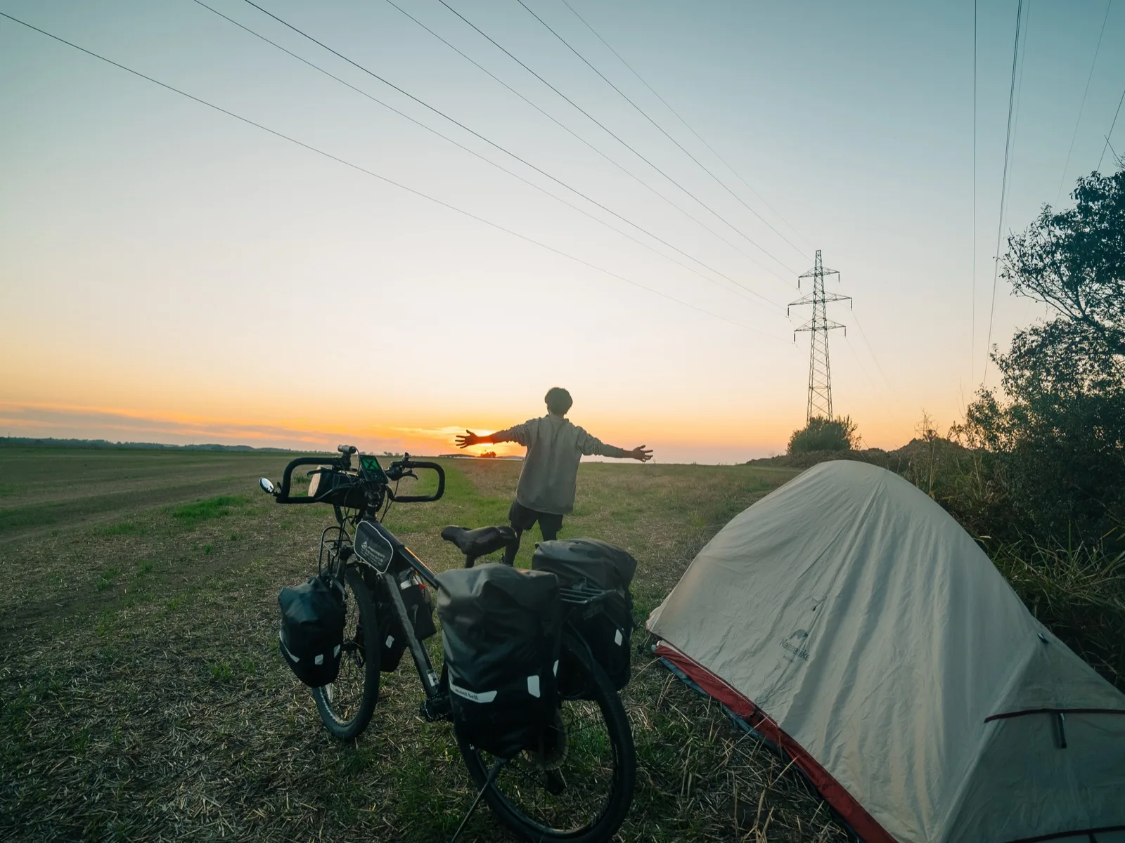 荷物を積んだ自転車とテント、夕日に向かって両手を広げる様子